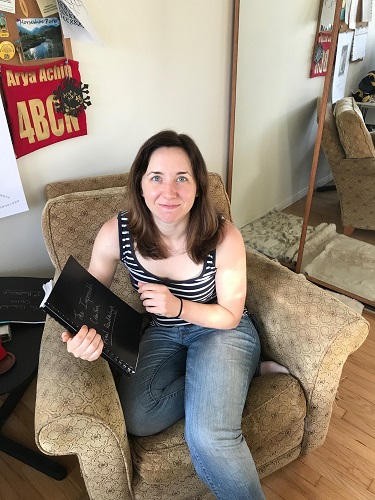 Author Betsy Flak holding a black bound notebook entitled 'The Impossible Creation: Final Readthrough, looking awkward.'